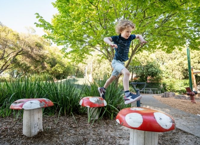 A young boy jumping between playground stumps that are painted to represent fairytale mushrooms in the daytime.