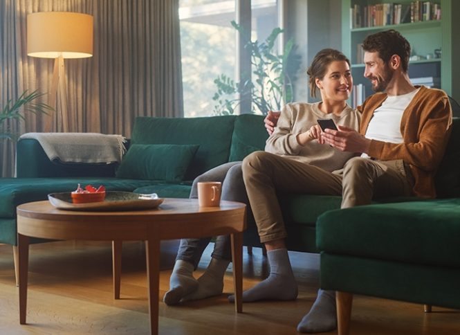 A man has his arm over a woman as they are seated on an emerald green couch, smiling at something on the man's phone.